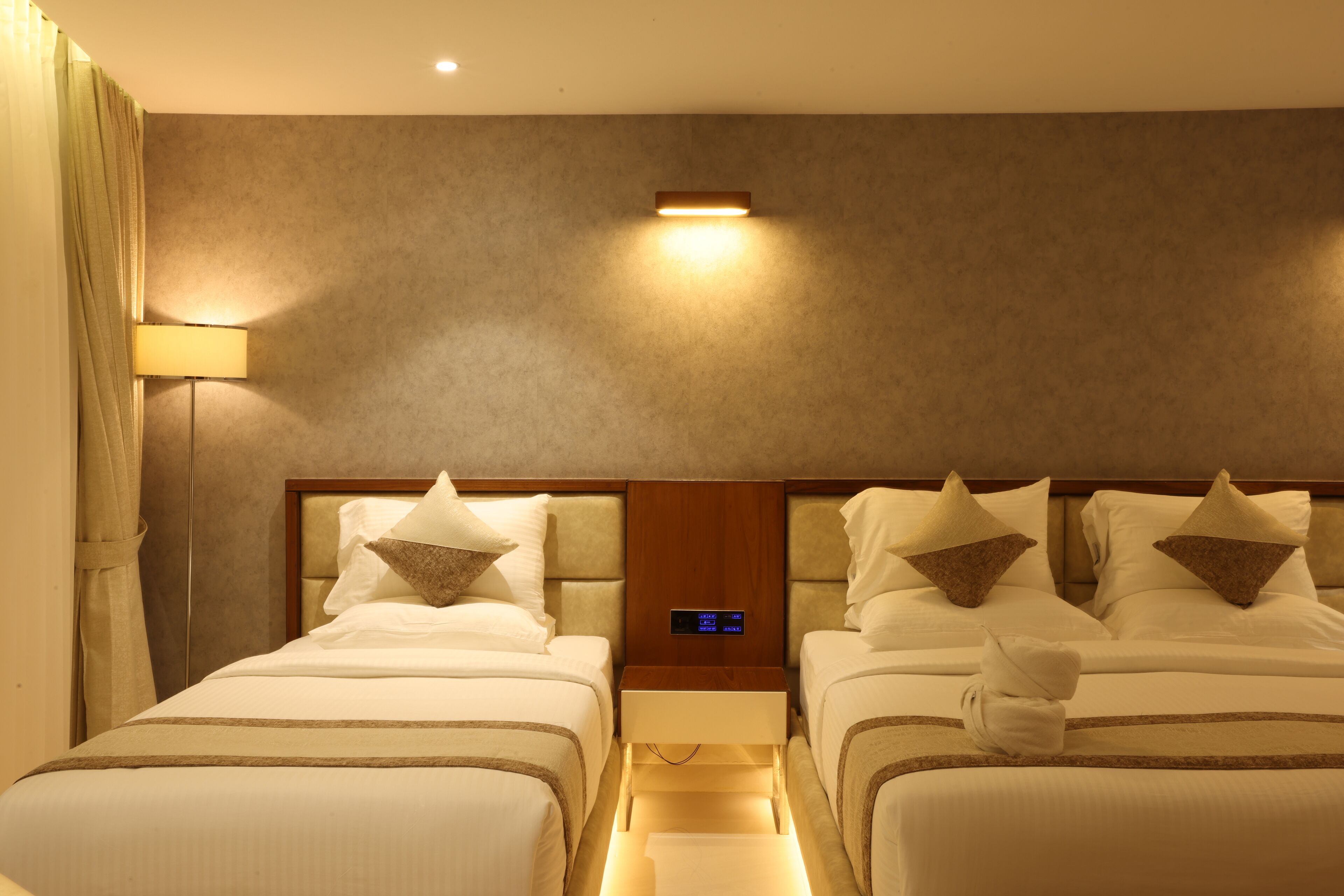 A hotel room with two beds, a nightstand, a digital clock, and a wall-mounted light.