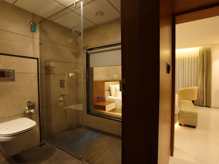 A modern hotel bathroom with a glass shower enclosure, a toilet, and a visible bed in the adjacent room.