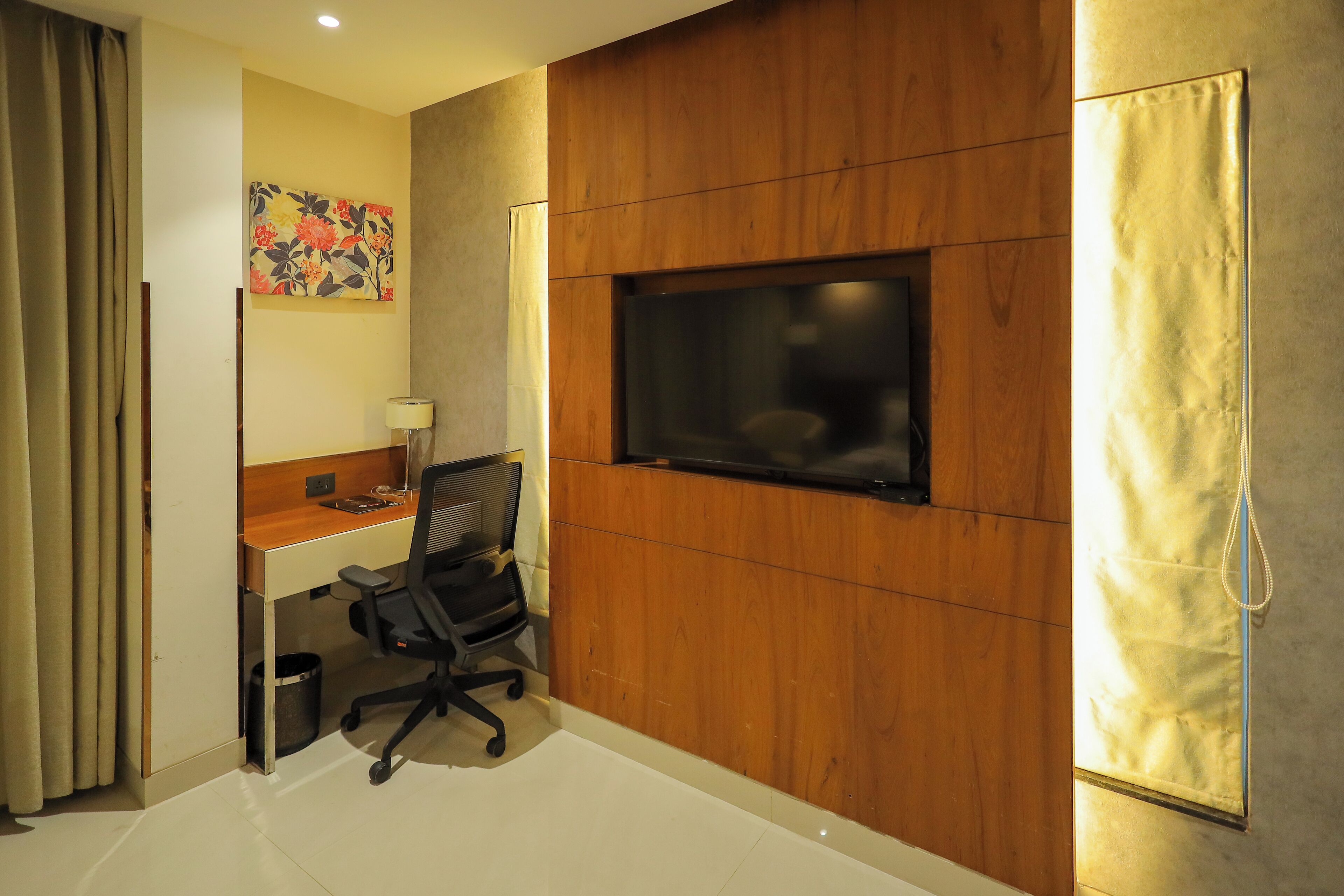 A hotel room with a wooden desk, a black office chair, a television mounted on the wall, and a curtain on the window.