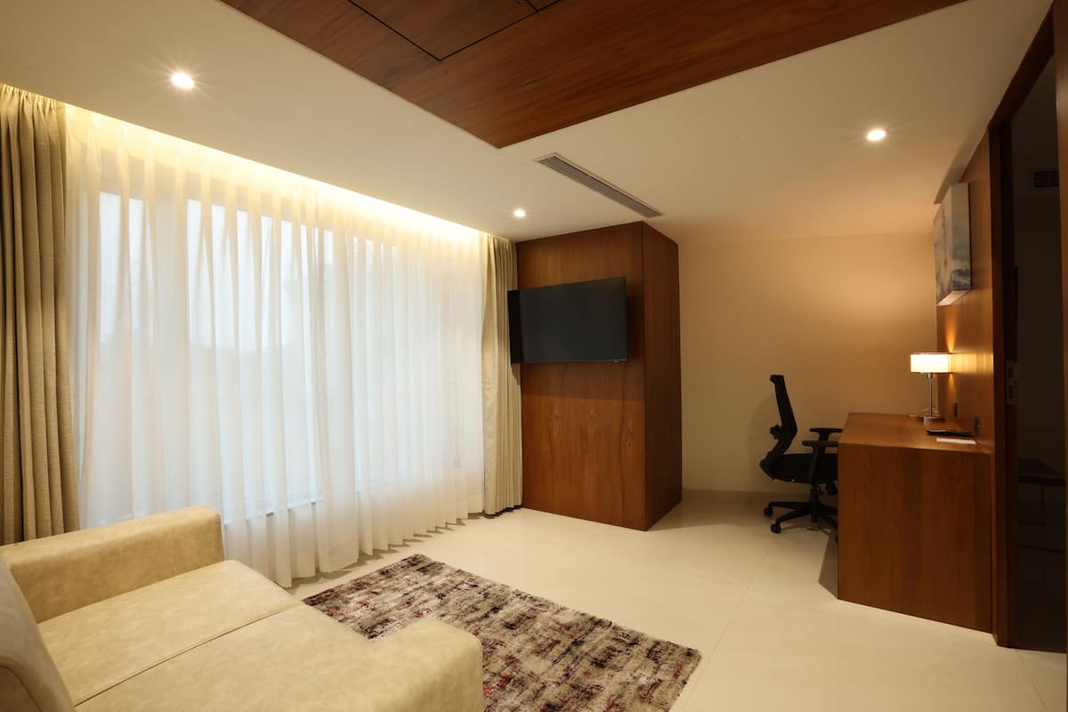 A modern hotel room with a sofa, a desk, a TV mounted on the wall, and a large window with curtains.