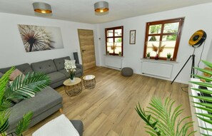 Un salon avec un canapé gris, un parquet en bois, une table basse ronde et une grande fenêtre ornée de plantes.