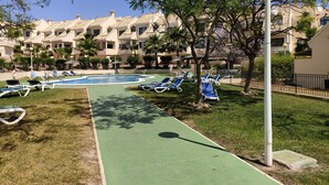 Un área de piscina con sillas de descanso y un sendero verde, rodeada de palmeras y un edificio con varios balcones.
