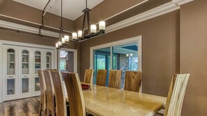 Uma sala de jantar com mesa e cadeiras de madeira, um candelabro e vista para o exterior através de uma porta de vidro.