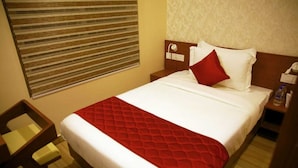 A hotel room with a bed, a nightstand, a bedside table, a red bedspread, and a window with blinds. - DCHANDELIER (Kanayannur)