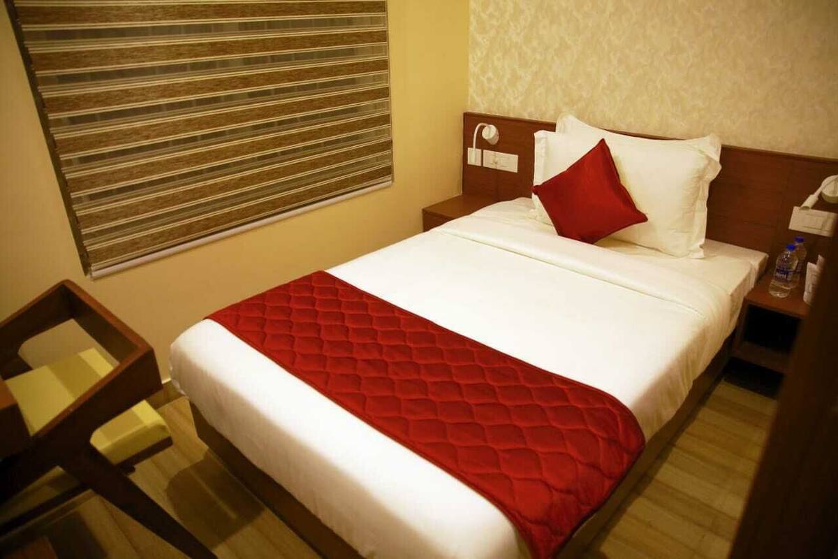 A hotel room with a bed, a nightstand, a bedside table, a red bedspread, and a window with blinds.