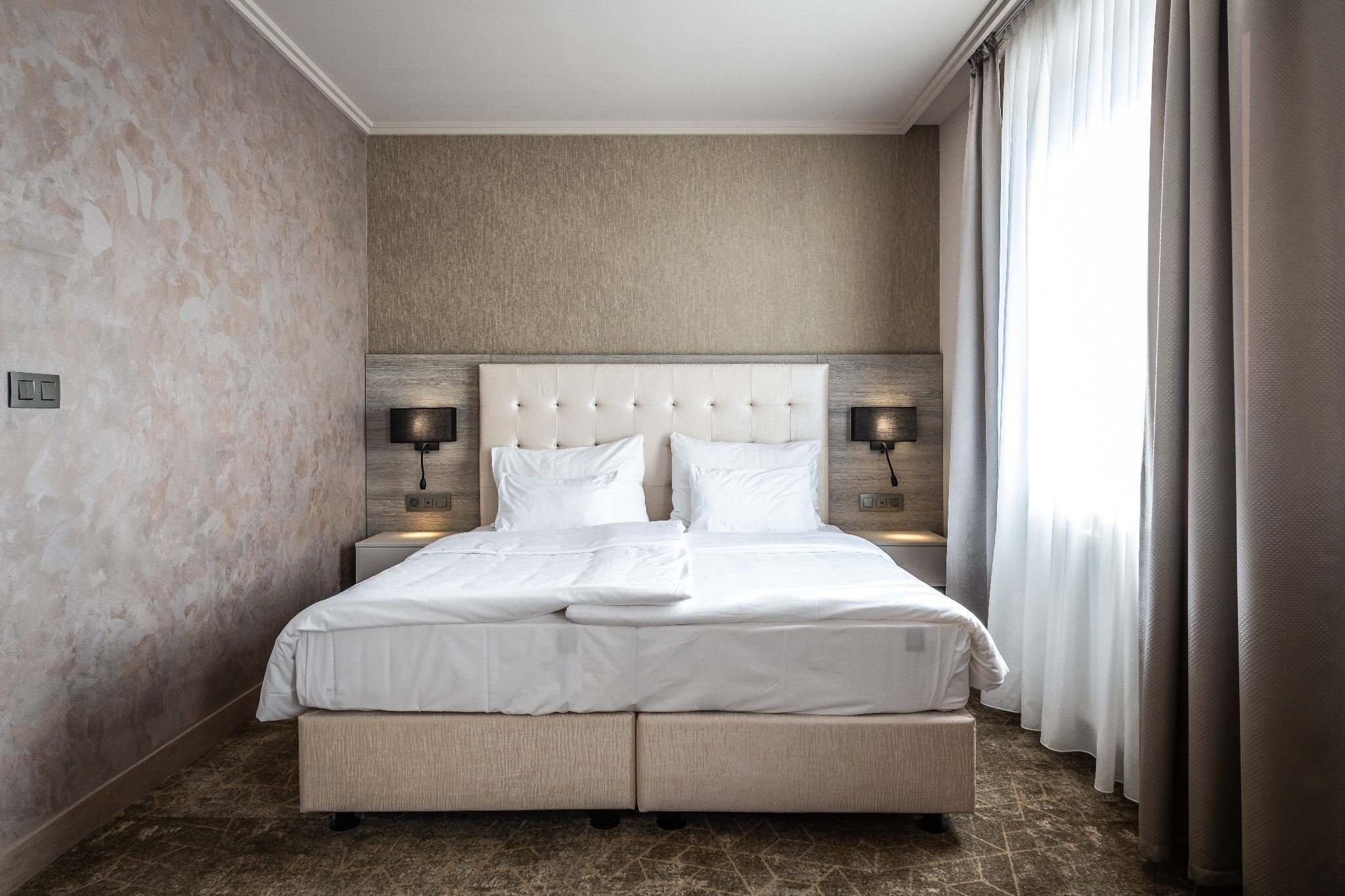 A hotel room with a large bed, two bedside tables with lamps, a headboard, and a view of the outdoors through a window with curtains.