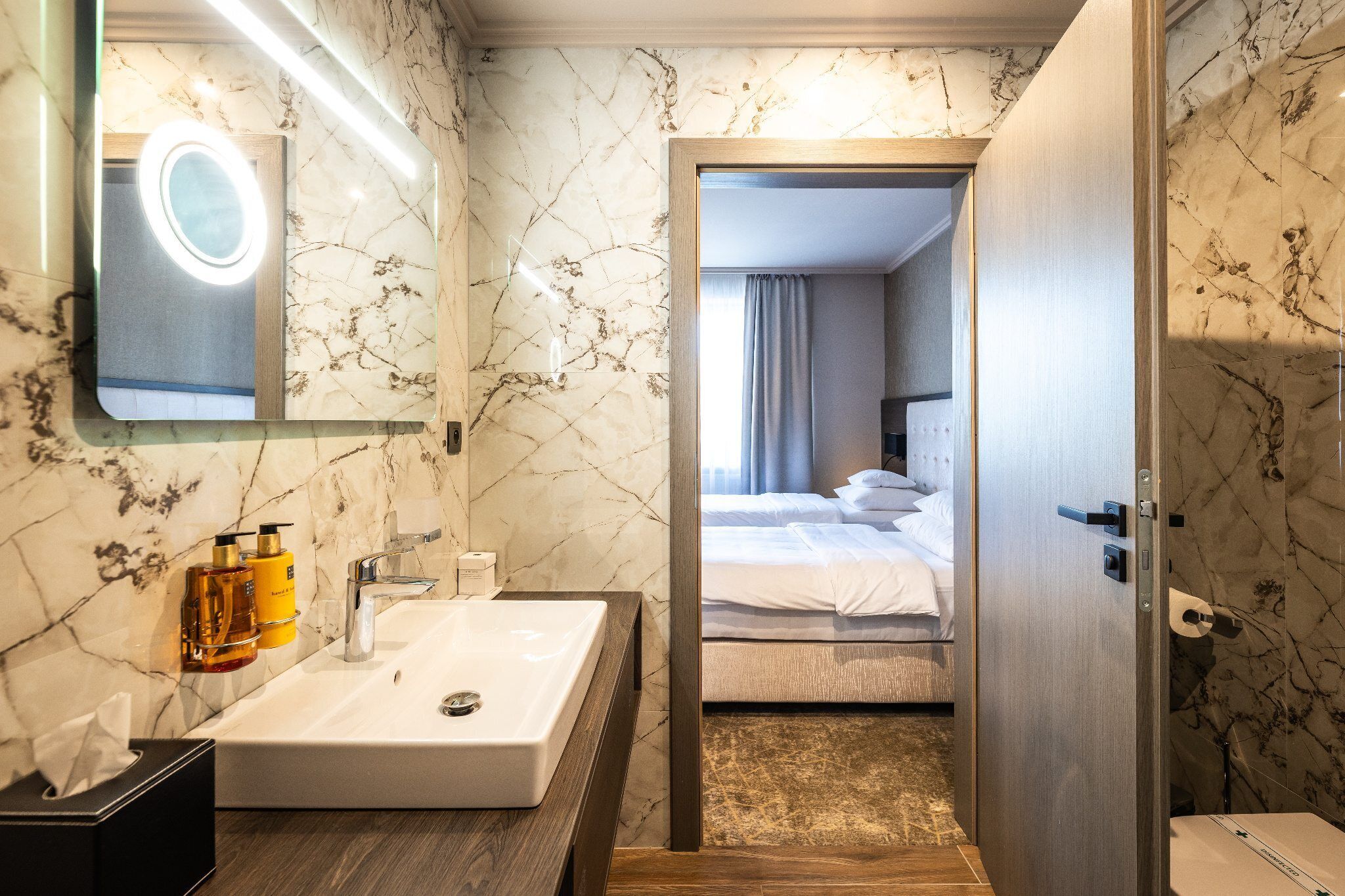 A hotel bathroom with a marble sink, a mirror with a light, and toiletries.
