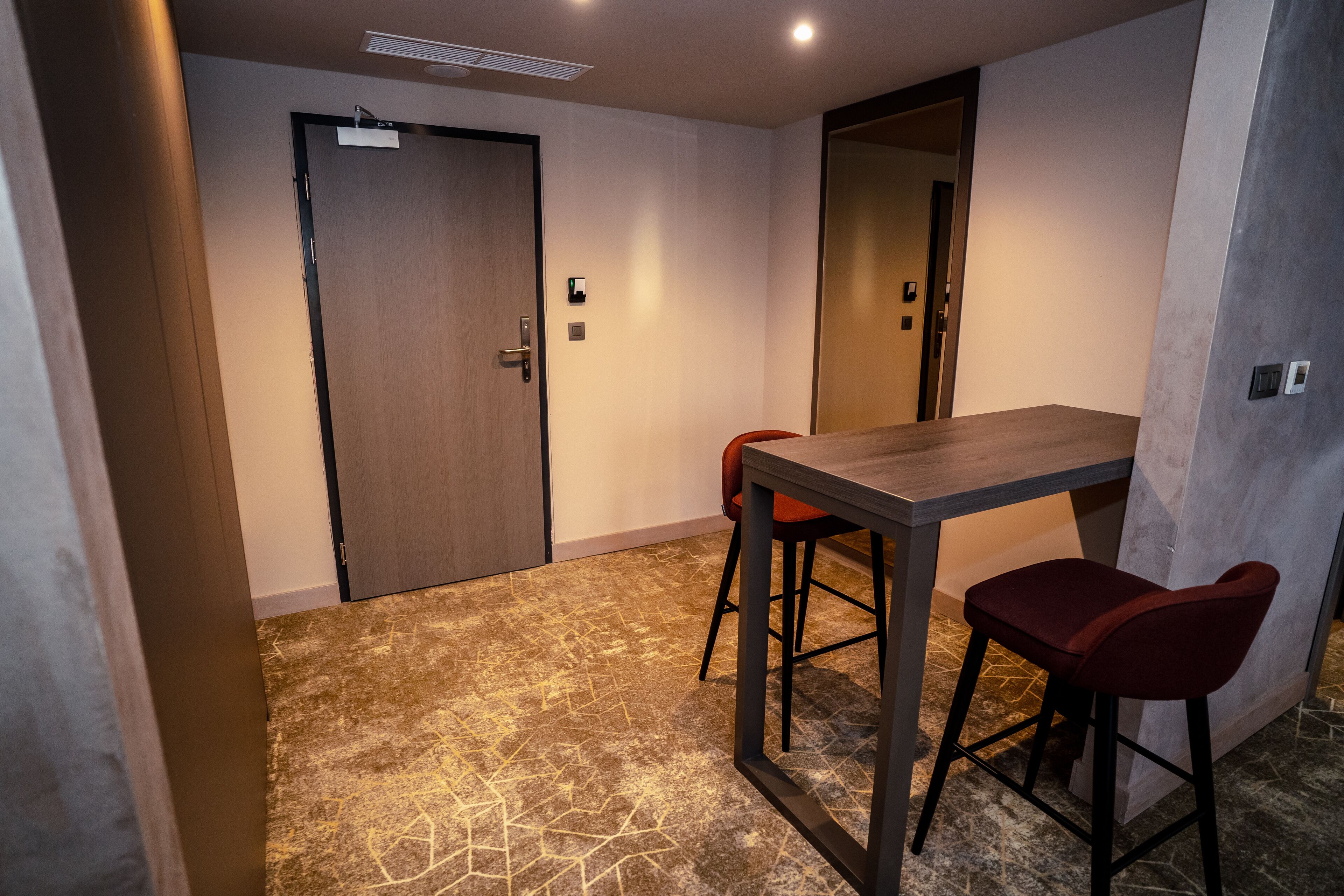 Una habitación con puerta de madera, una mesa pequeña con dos sillas rojas y una alfombra estampada.