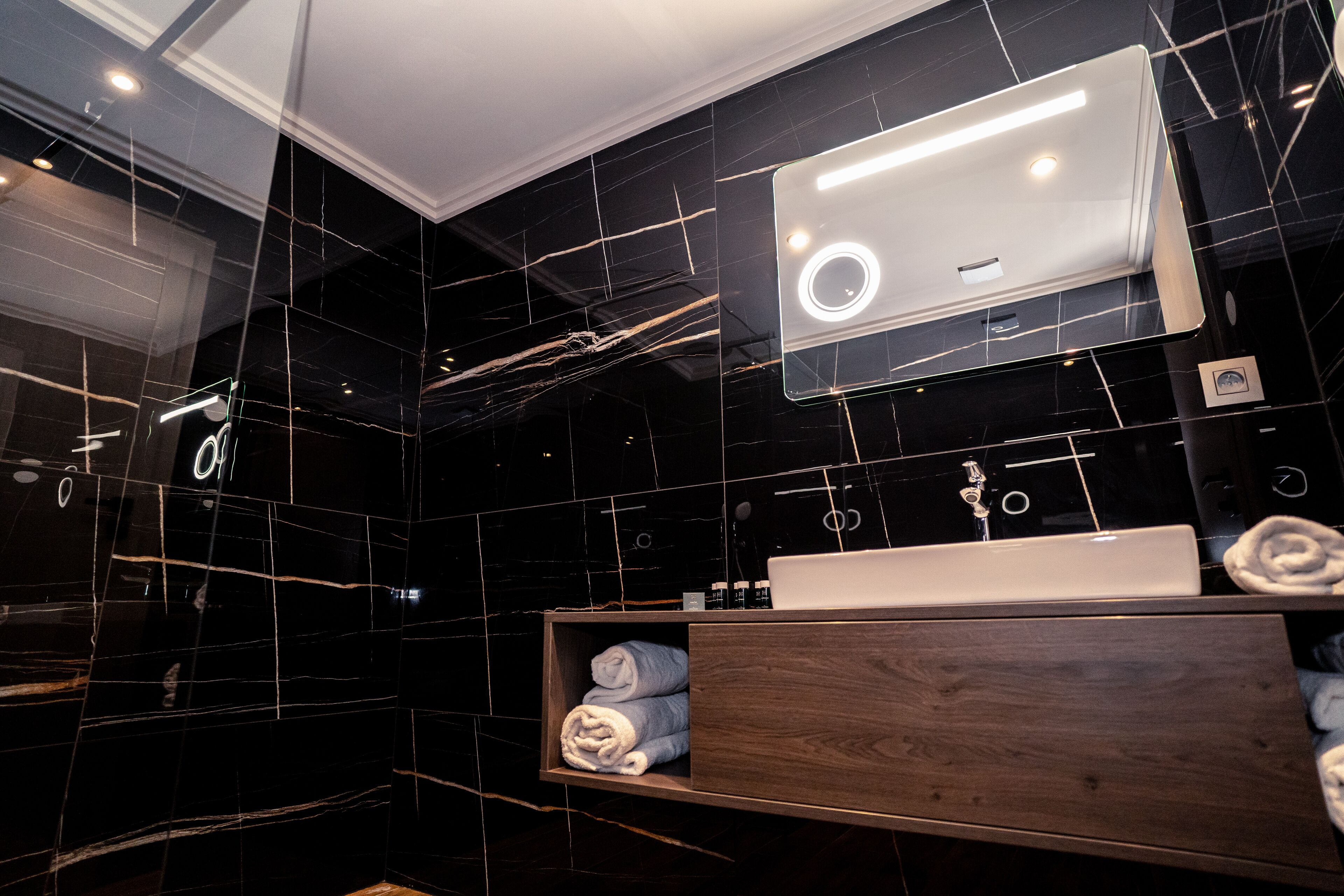 A modern bathroom with a dark marble backsplash, a wooden vanity, a white sink, and a mirror with integrated lighting.