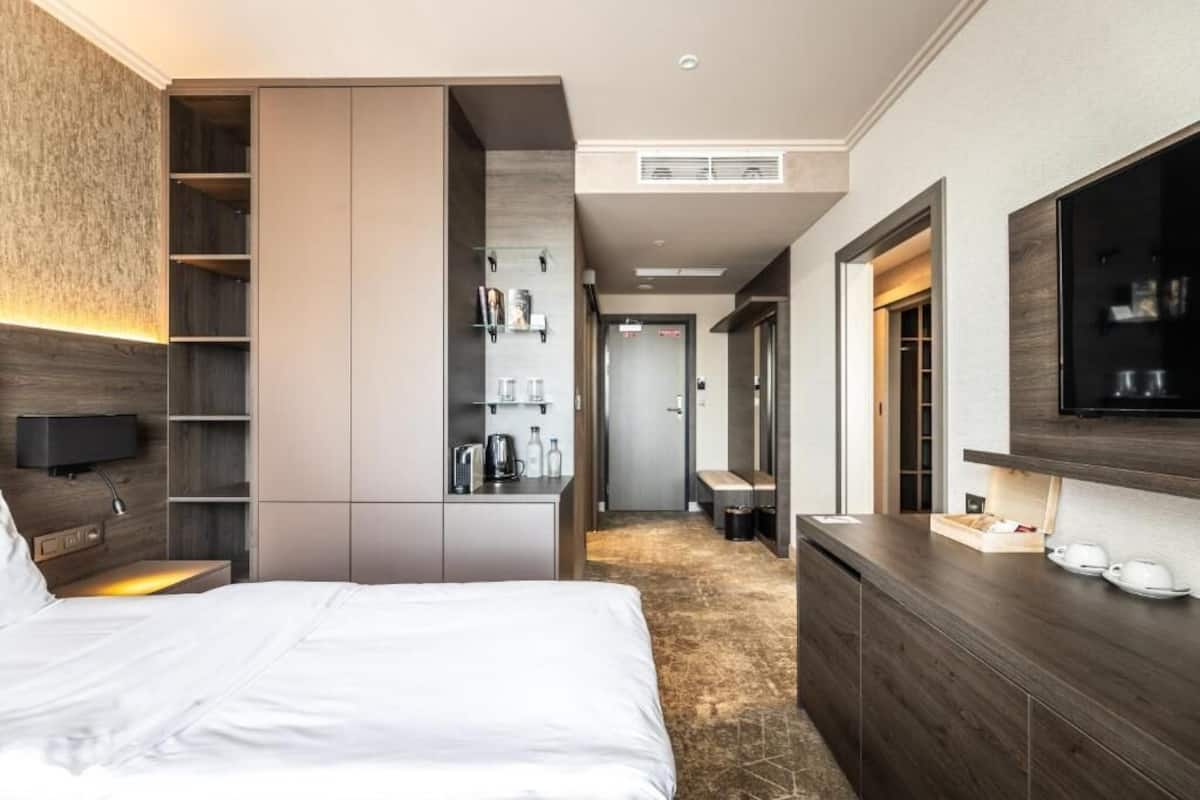 A modern hotel room with a large bed, built-in wardrobe, a kitchenette, and a TV mounted on the wall.