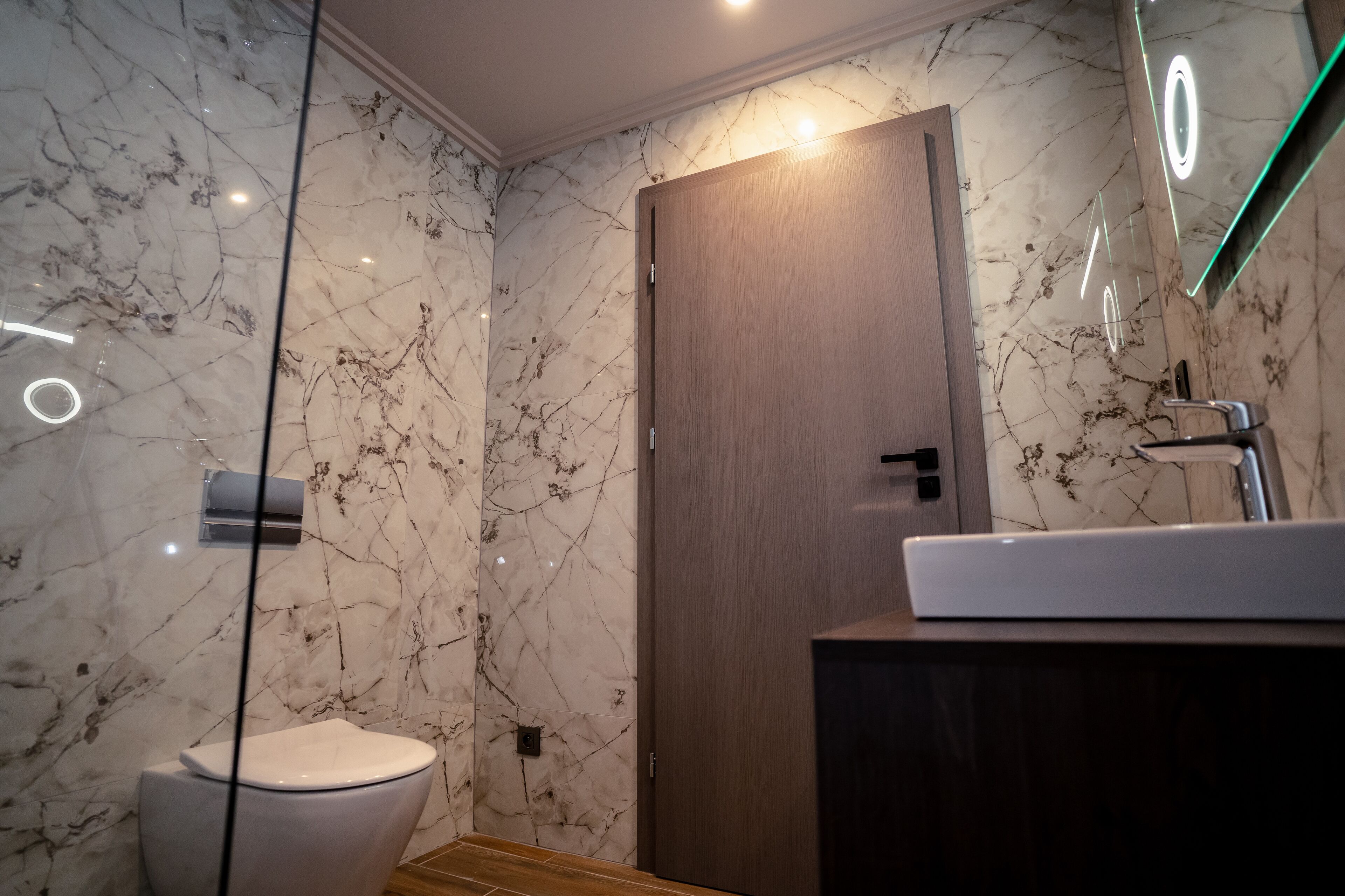 Un baño moderno con pared de mármol, puerta de madera, inodoro blanco y tocador de madera con espejo.
