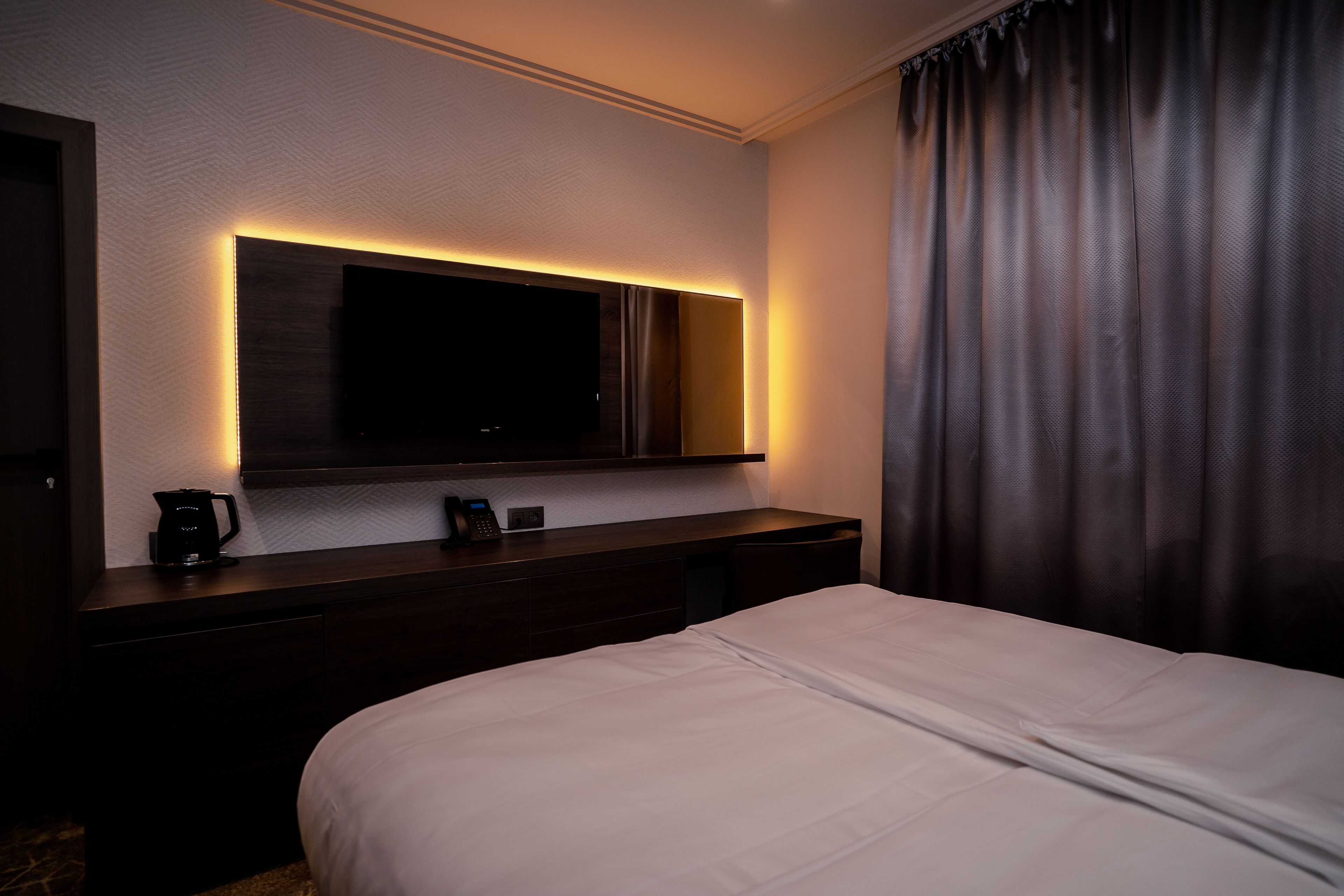 A hotel room with a large bed, a flat-screen TV mounted on a wall, a bedside table with a coffee maker, and a curtain on the window.
