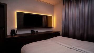 Habitación de hotel con una cama grande, un televisor de pantalla plana montado en la pared, una mesita de noche con cafetera y una cortina en la ventana.