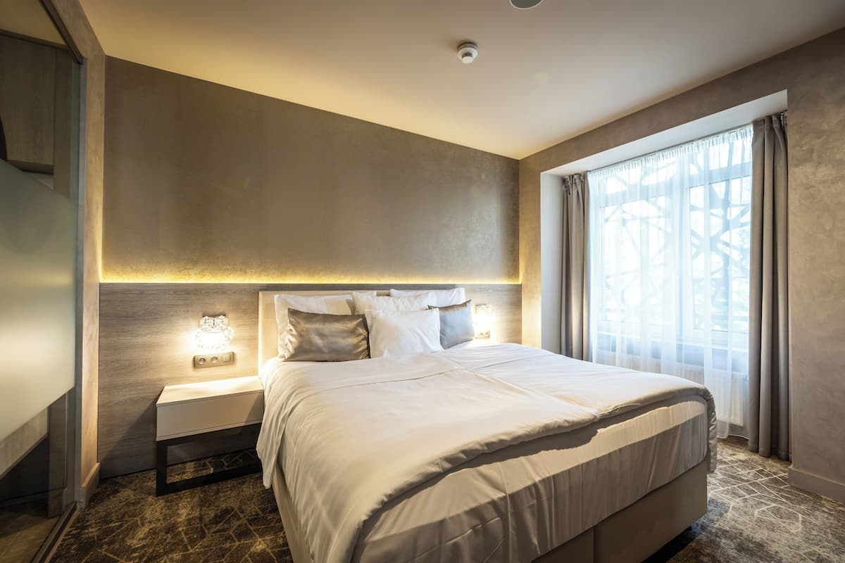 A modern hotel room with a large bed, a nightstand with a lamp, and a window with sheer curtains.