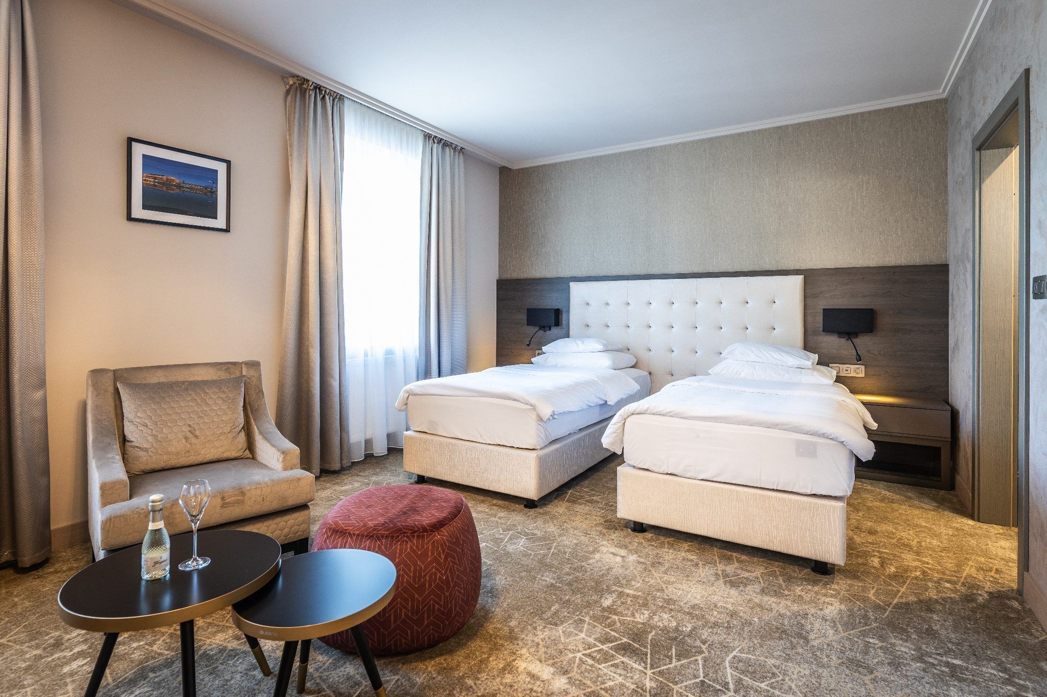 A hotel room with a bed, a chair, a small table, and a red ottoman.
