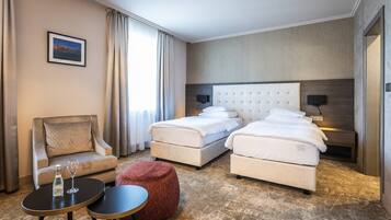 A hotel room with a bed, a chair, a small table, and a red ottoman.