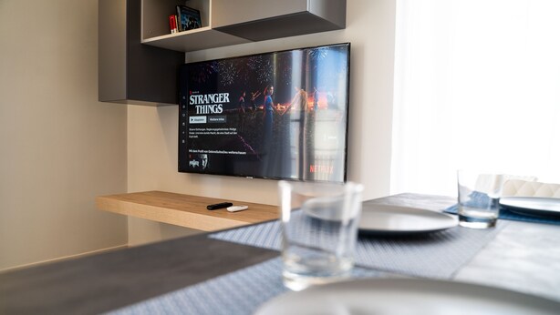 Une chambre d’hôtel moderne équipée d’un téléviseur à écran plat diffusant la série « Stranger Things », d’une table de chevet en bois avec une télécommande, d’un verre d’eau et d’une table à manger avec des assiettes et des couverts.