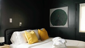 Een net opgemaakt bed met witte en gele kussens, een opgevouwen handdoek en een ingelijst kunstwerk aan de muur.