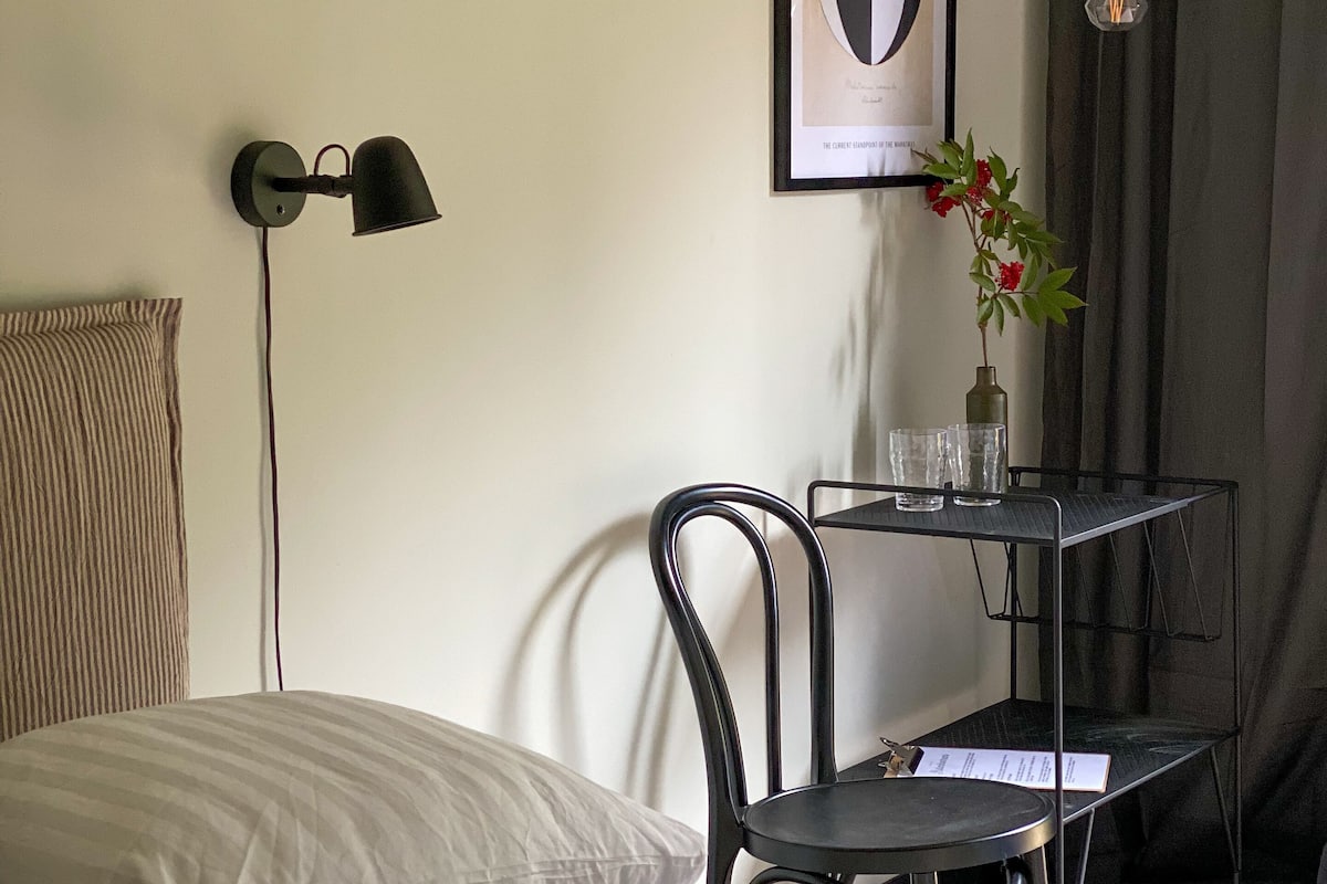 Quarto bem arrumado com cama, uma cadeira, uma mesinha com copos, um quadro na parede e um abajur.