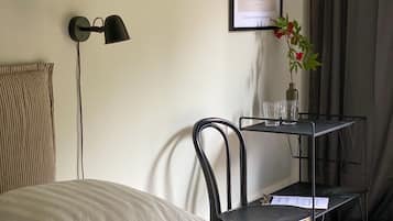 Quarto bem arrumado com cama, uma cadeira, uma mesinha com copos, um quadro na parede e um abajur.