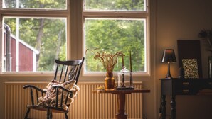A room with a rocking chair, a table with a vase and candle, and a view of greenery outside. - Rallarhustruns (Hassleholm)