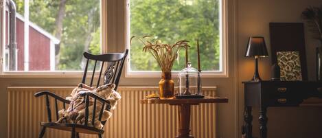Una habitación con una mecedora, una mesa con un jarrón y una vela, y vistas a zonas verdes al exterior.