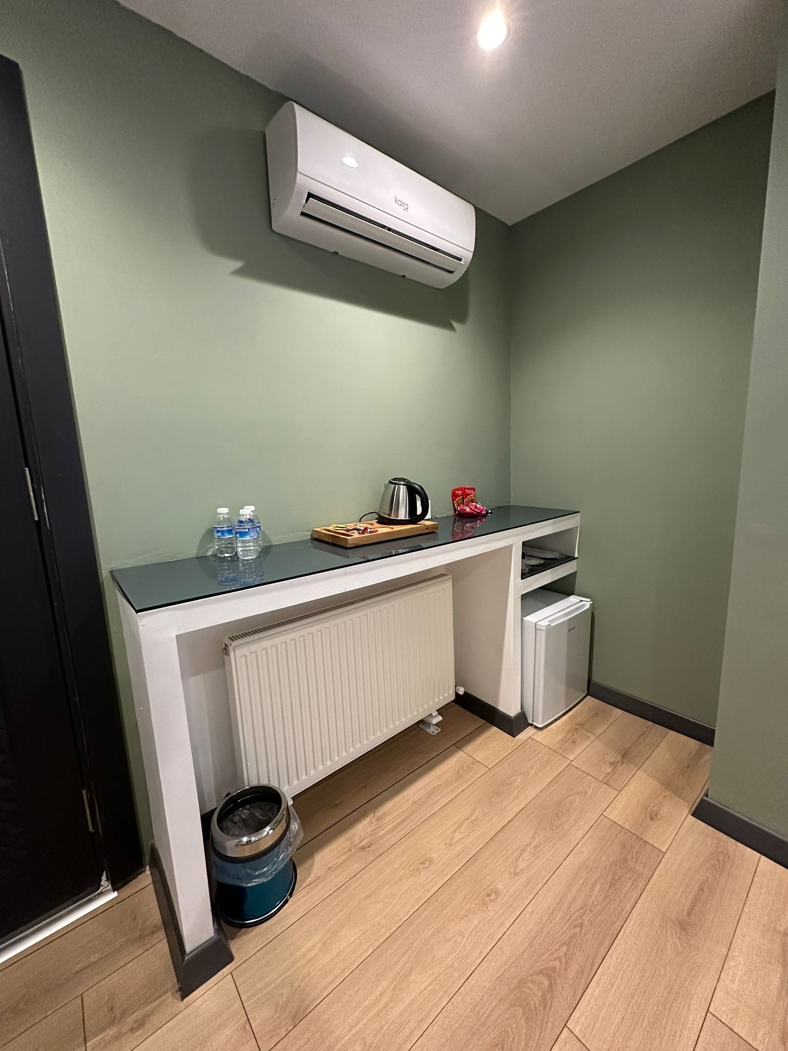 Uma cozinha compacta com bancada branca, chaleira, mini-geladeira e radiador.
