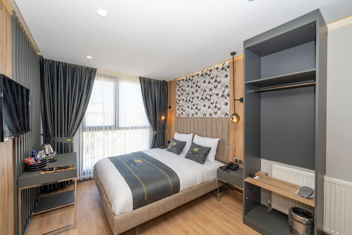 A modern hotel room with a large bed, a desk with a flat-screen TV, a wardrobe, and a window with curtains.