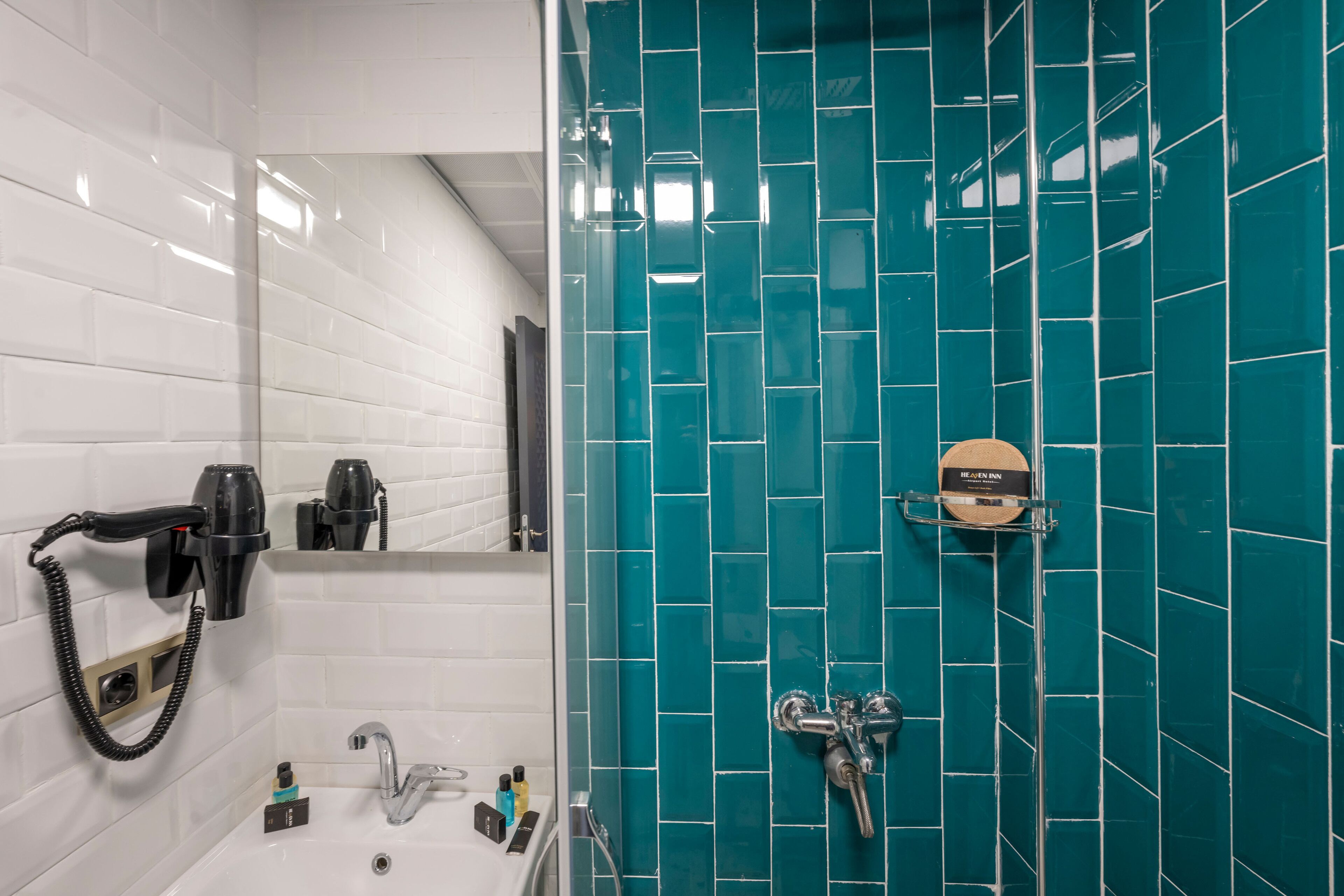 A bathroom with white tiled walls, a sink, a mirror, a hairdryer, and a shower with a teal tile backsplash.