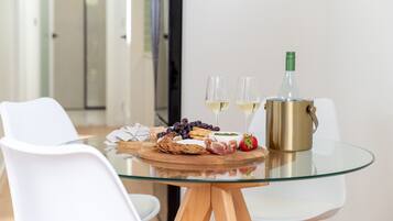A glass table with a bottle of white wine, two glasses, a wooden board with snacks, and a white chair.