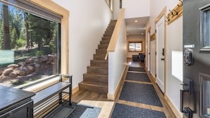 A modern interior with a wooden staircase, a view of a forest through a large window, and a door with a keypad lock. - Pine Cone Place_Brand New Build, EV Charger, Hydronic Heat, Steam Shower, 500G Wifi, Arcade & PS4 (Truckee)