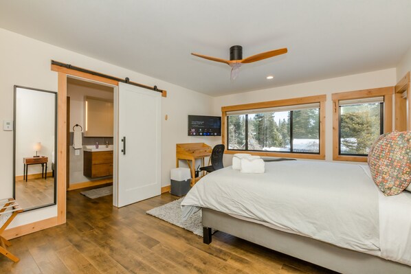 A bedroom with a large bed, a desk, and a chair, a ceiling fan, and a view of trees through the windows.