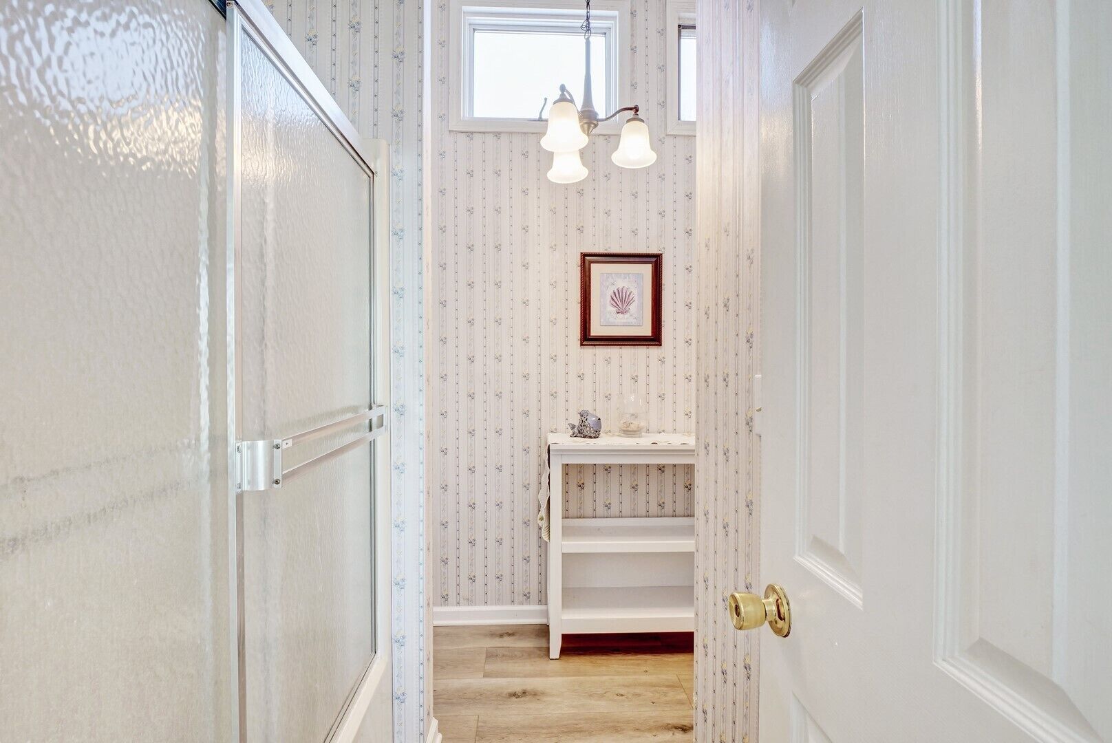 A narrow hallway with a wooden floor, a white door on the right, and a white door on the left. There is a small shelf with decorative items and a framed picture on the wall.