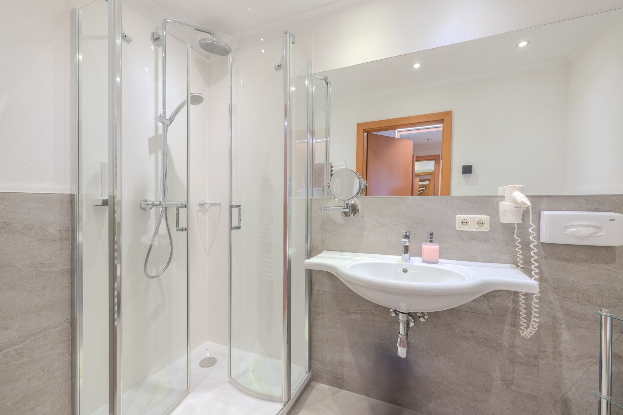 Un baño moderno con ducha de vidrio, un lavamanos blanco y un dispensador de jabón rosa, y un secador de manos.