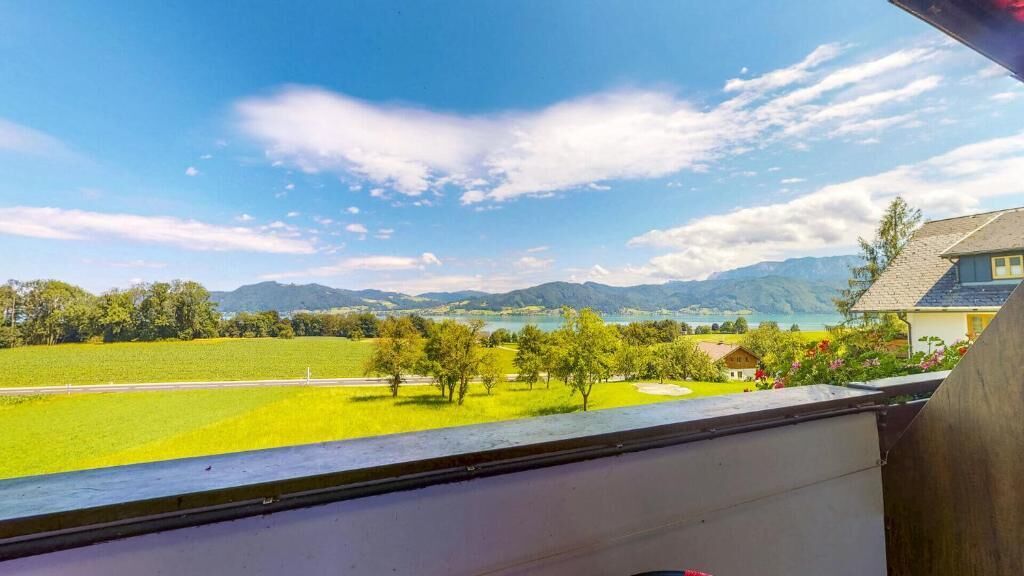 A view from a window overlooking a green field, a small house, and distant mountains under a clear sky.