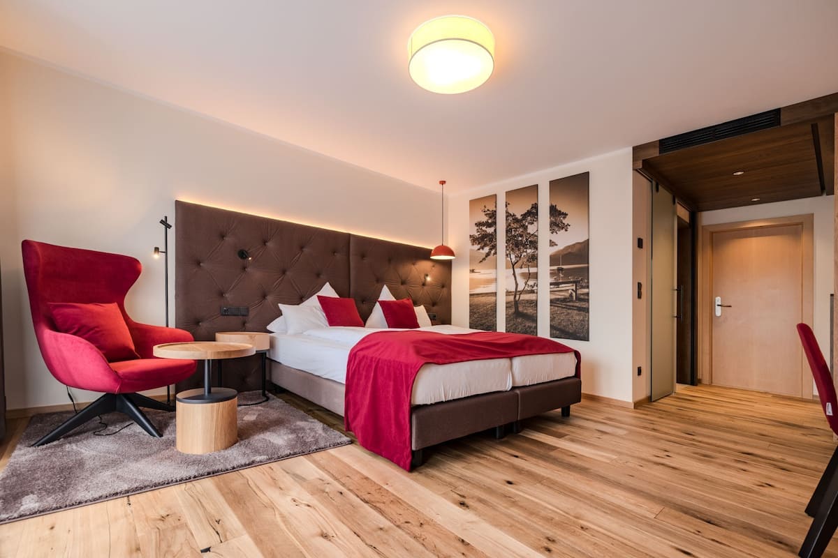 Une chambre moderne avec une tête de lit capitonnée, une chaise rouge, une table ronde en bois et un lit avec une literie rouge.