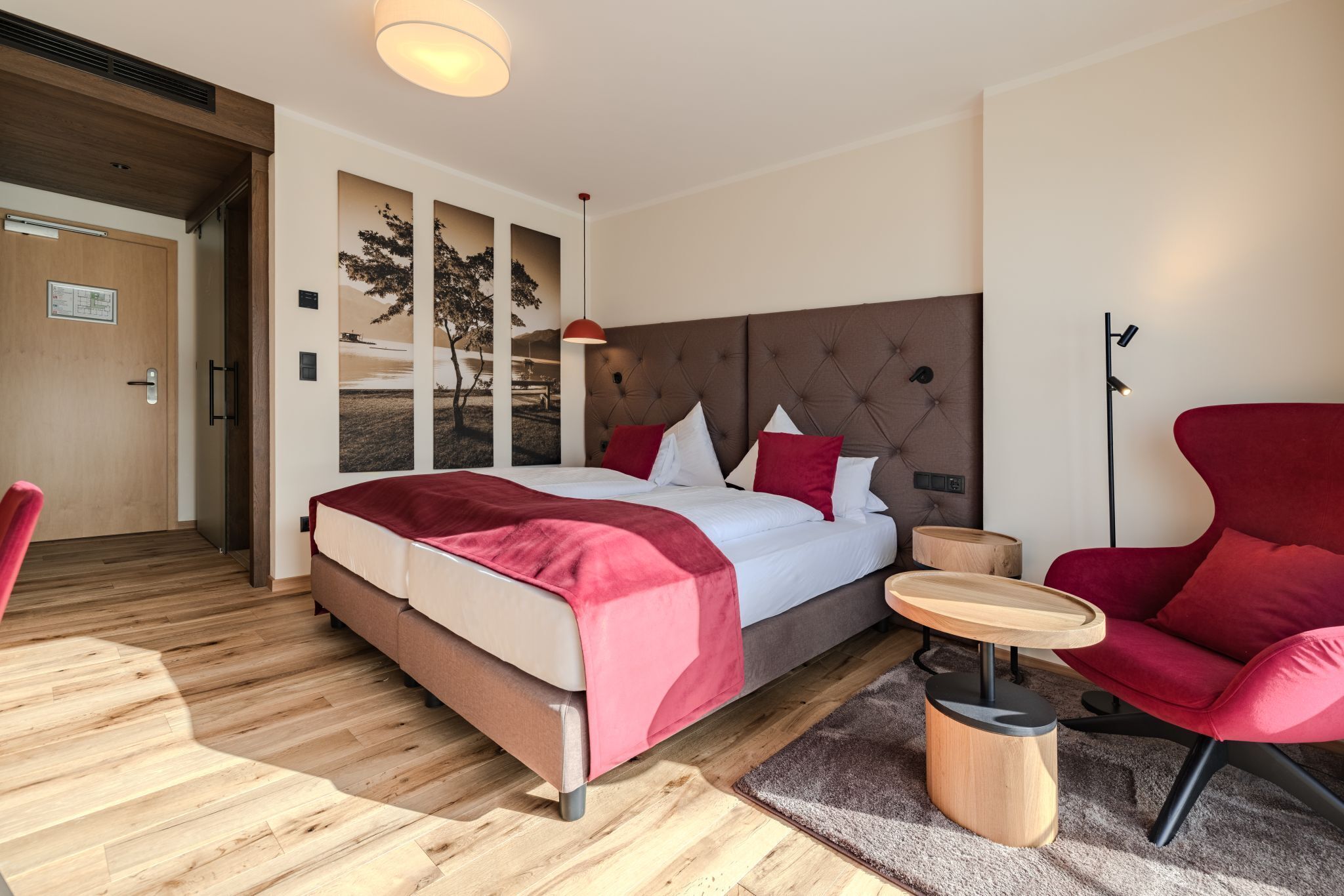 A modern hotel room with a large bed, a red armchair, a small round table, and a wooden floor.