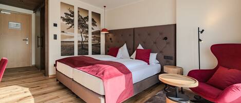 A modern hotel room with a large bed, a red armchair, a small round table, and a wooden floor.
