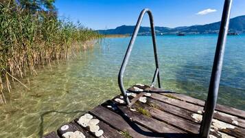 A wooden dock extending into clear water with a metal railing, surrounded by reeds and distant mountains.