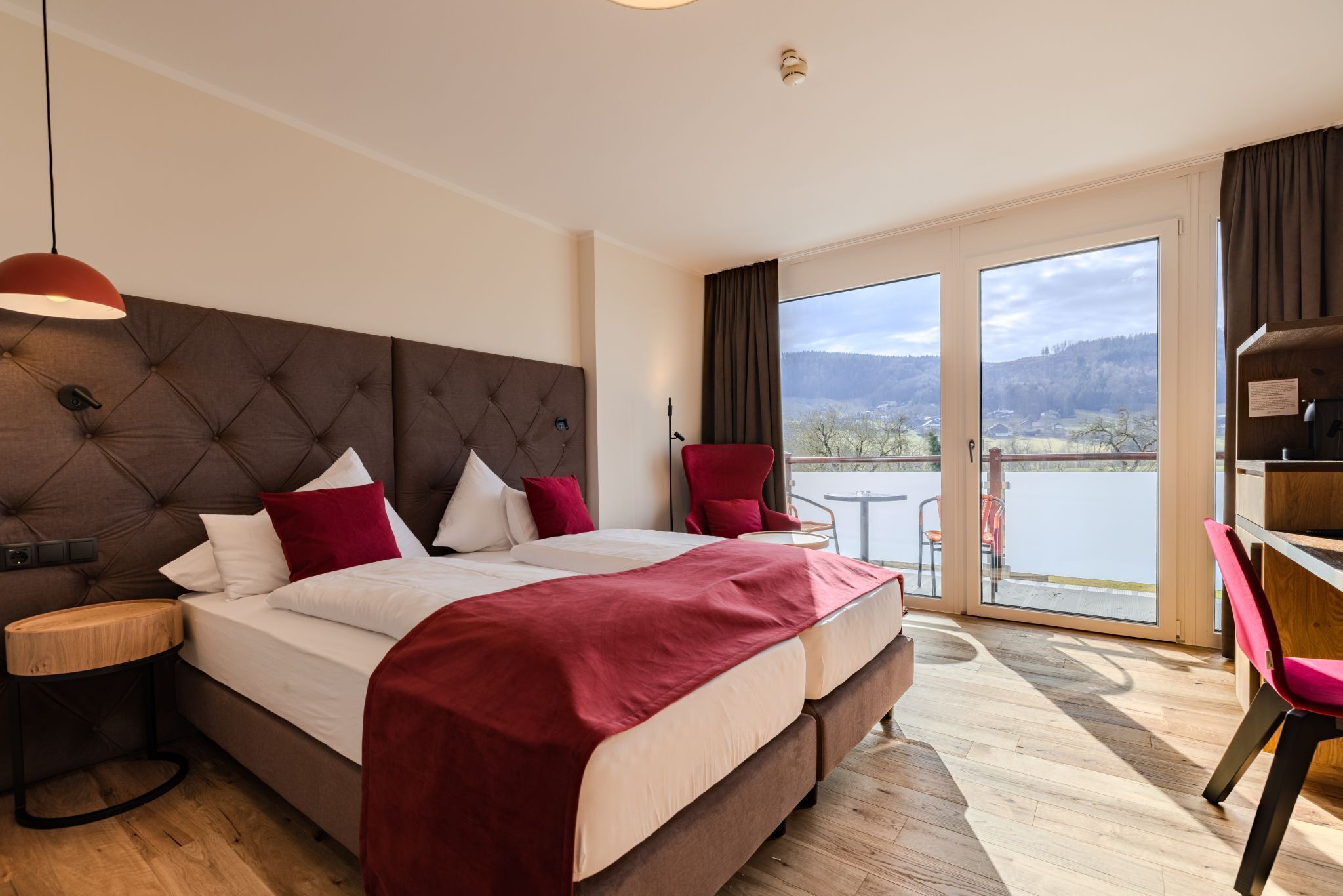 A modern hotel room with a large bed, a red chair, a desk, and a view of a lake.