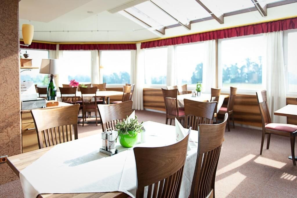 A dining area with multiple tables and chairs, large windows with curtains, and a view of greenery outside.