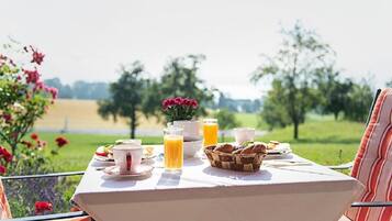 A table set outdoors with breakfast items, a basket of bread, and a view of a green landscape.