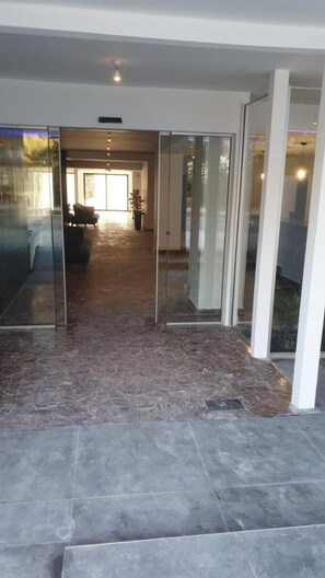 Un pasillo con una puerta de cristal que da a una sala de estar, suelo de baldosas y una lámpara en el techo.
