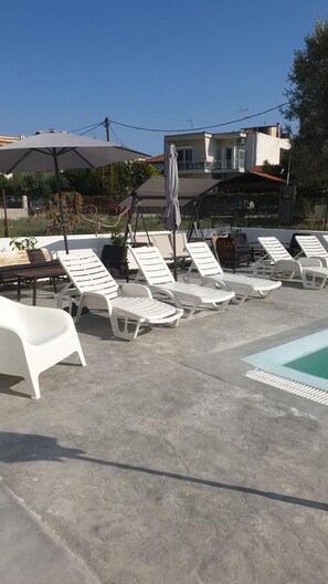A poolside area with white lounge chairs and umbrellas, a concrete floor, and a building in the background. - Evita Blue Hotel (Xylokastro-Evrostina)