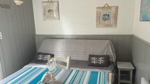 A small, neatly arranged room with a bed, a table with a striped tablecloth, and two framed pictures on the wall. - Cottage Les Roses de mer (Sainte-Marie-de-Ré)