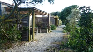 A gravel path leading to a wooden structure with a table and chairs, surrounded by dense greenery and trees. - Cottage Les Roses de mer (Sainte-Marie-de-Ré)