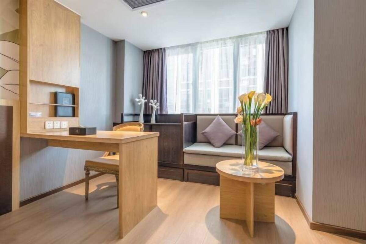 Habitació d'hotel moderna amb un escriptori de fusta, una taula rodona amb un ram i un sofà amb coixins, i una finestra amb cortines.