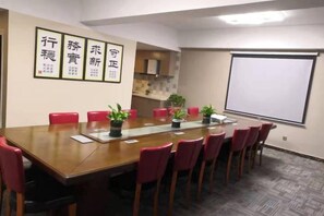 Un salón de conferencias con una larga mesa, sillas rojas, una pantalla de proyector y una pared con textos enmarcados.