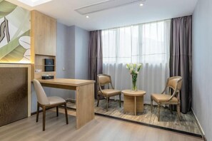 Une chambre d’hôtel moderne avec un bureau en bois, deux chaises, une petite table ronde ornée d’un vase de fleurs et de grandes fenêtres avec des rideaux.