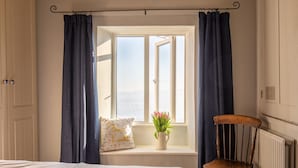 A bedroom with a window, a wooden chair, a bedside table with a plant, and a pillow with a floral pattern. - Swift Cottage, Blockley - sleeps 4 guests in 2 bedrooms (Blockley)